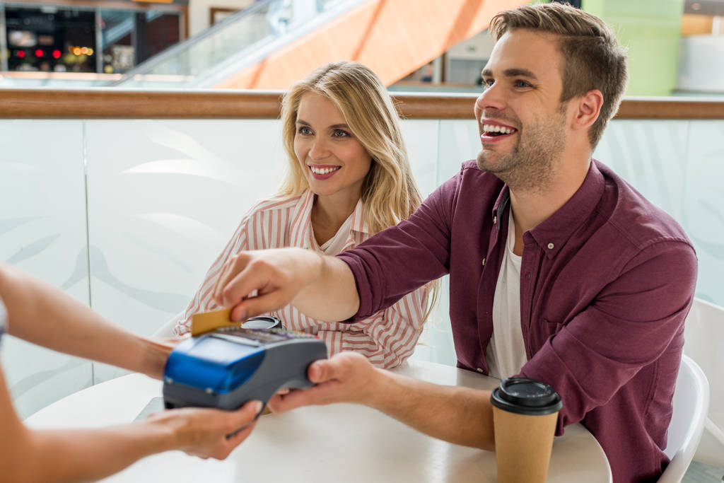 χαμογελαστός νεαρός άνδρας που πληρώνουν με πιστωτική κάρτα στο τερματικό, ενώ τη φίλη που κάθονται κοντά στο τραπέζι με φλιτζάνια καφέ σε καφετέρια  - Φωτογραφία, εικόνα