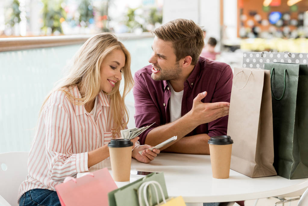 mosolygó fiatal nő számolás készpénzre, ideges barátja kávéscsészét és papír táskák cae asztalnál ül míg vásárlás után  - Fotó, kép