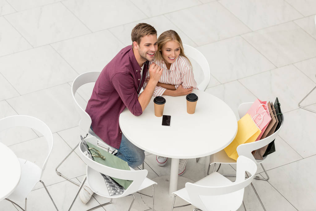 μεγάλη γωνία θέα ζευγάρι με τσάντες αγορών που κάθεται στο τραπέζι με ποτήρια μιας χρήσης και smartphone με κενή οθόνη στην καφετέρια  - Φωτογραφία, εικόνα