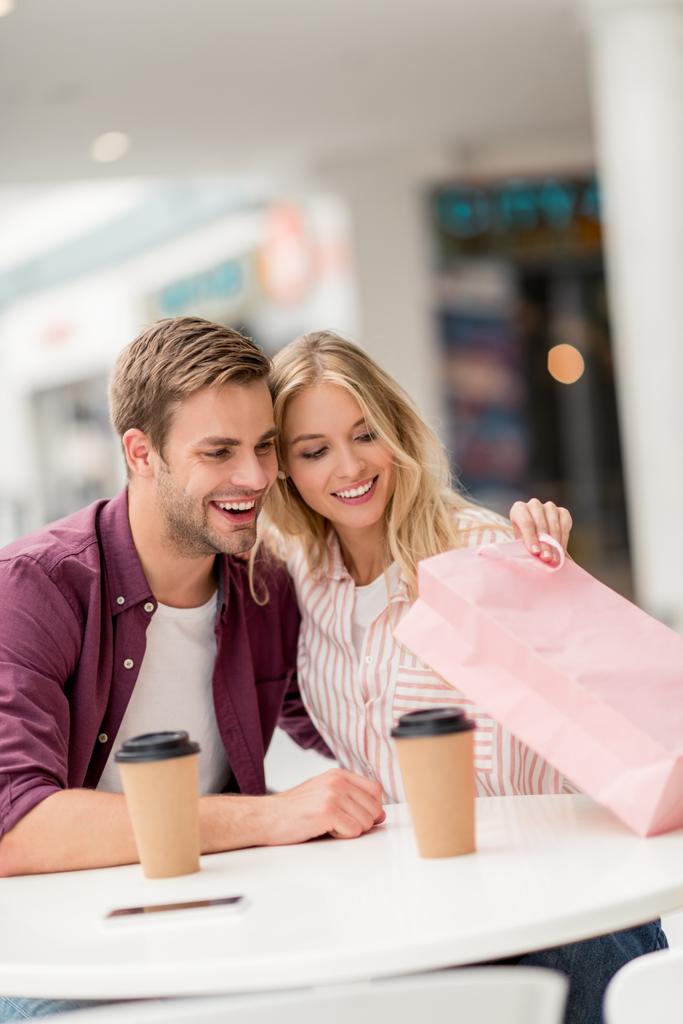 selectieve aandacht van jonge vrouw op zoek naar boodschappentas met vriendje aan tafel met koffie kopjes in café  - Foto, afbeelding