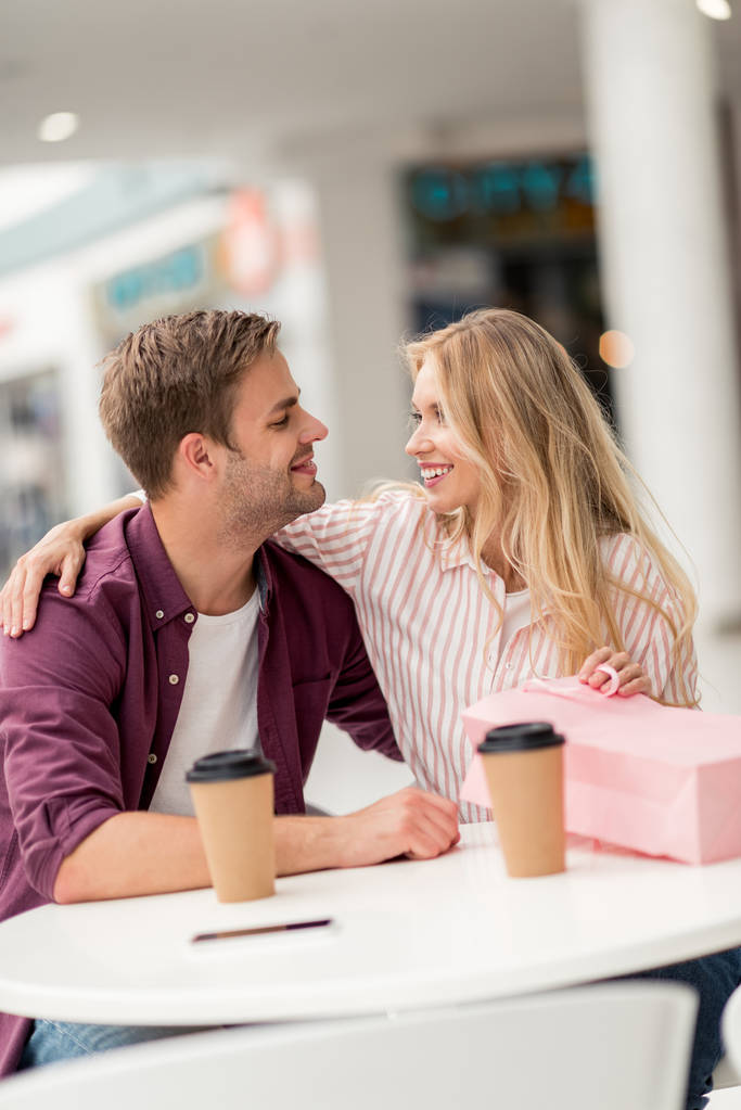 sonriente pareja con bolsa de compras mirándose a la mesa con tazas desechables en la cafetería
 - Foto, Imagen