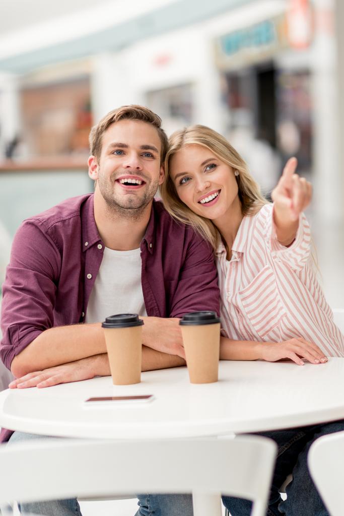 attractive young woman pointing by finger to smiling boyfriend at table with disposable cups of coffee in cafe - Photo, Image