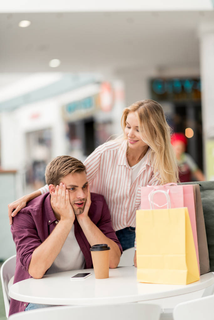 shocked young man looking at shopping bags at table with paper cup of coffee while his girlfriend standing near in cafe  - Photo, Image