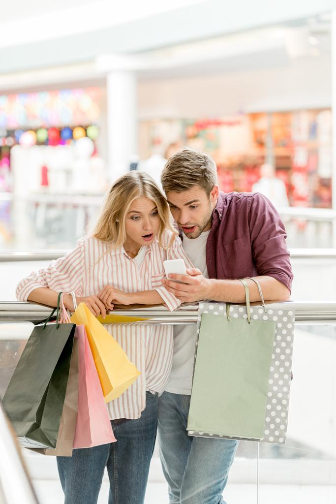 casal chocado de compradores com sacos de papel olhando para smartphone no shopping
  - Foto, Imagem