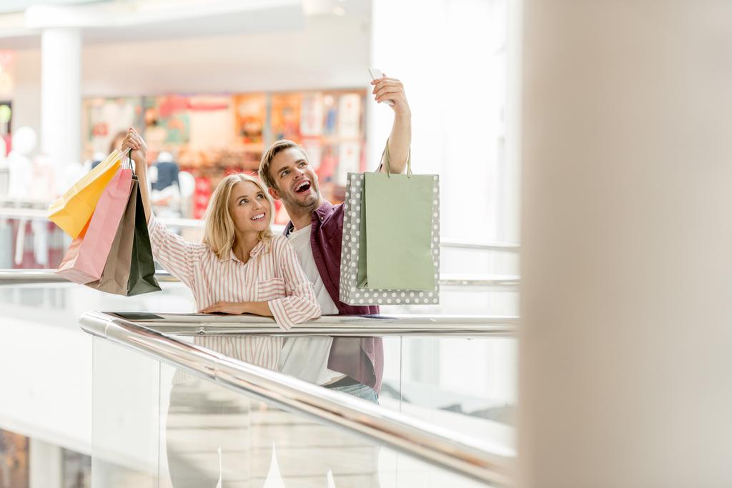 enfoque selectivo de joven sonriente pareja de compradores con tomar selfie con bolsas de compras en el centro comercial
  - Foto, imagen