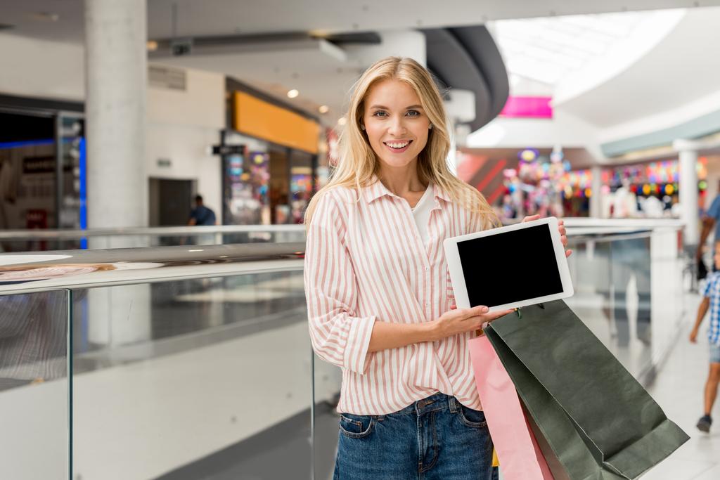 junge lächelnde Frau mit Einkaufstaschen zeigt digitales Tablet mit leerem Bildschirm im Einkaufszentrum  - Foto, Bild