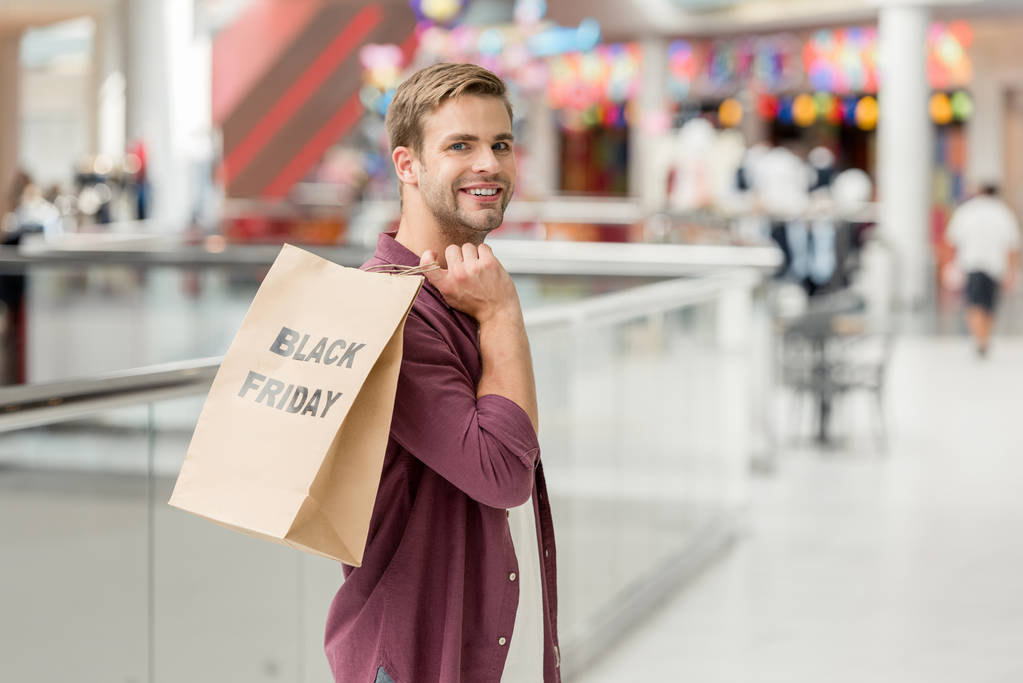 νέοι χαμογελαστός άνθρωπος που κρατά χαρτί με επιγραφή Μαύρη Παρασκευή και βλέπουν φωτογραφική μηχανή στο εμπορικό κέντρο  - Φωτογραφία, εικόνα
