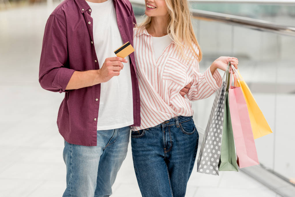 částečný pohled člověka ukazuje kreditní kartu a jeho přítelkyně, stojící poblíž nákupní tašky v obchoďáku  - Fotografie, Obrázek
