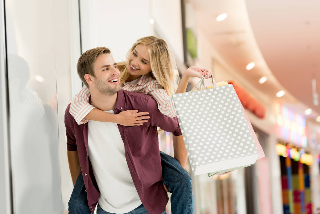 若い男がモールで買い物袋のガール フレンドに与えるおんぶ  - 写真・画像