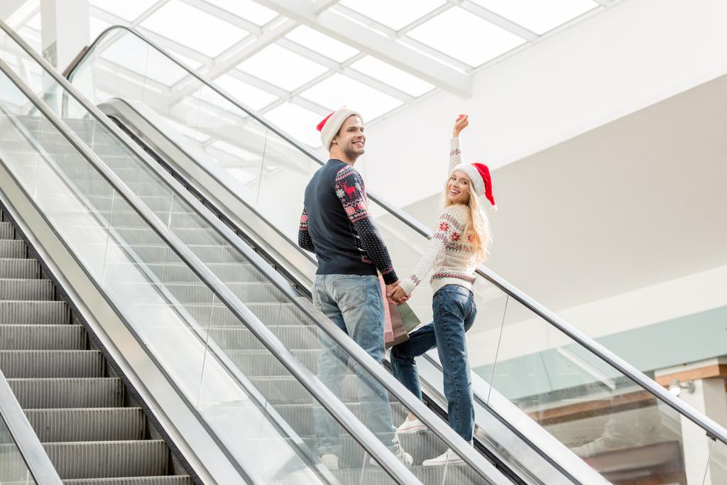 jeune femme en chapeau de Noël avec les bras levés regardant la caméra tandis que son petit ami debout près de l'escalator
  - Photo, image