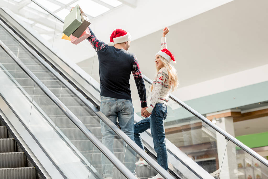 vista posteriore di coppia di acquirenti in cappelli natalizi con braccia sollevate in possesso di sacchetti di carta sulla scala mobile
  - Foto, immagini