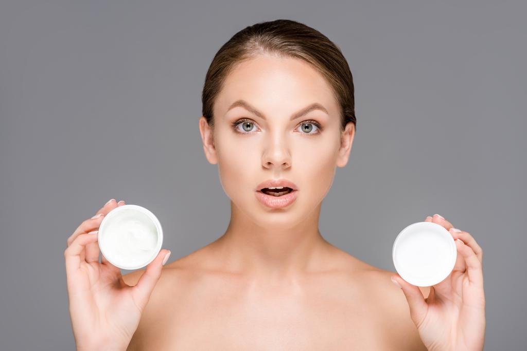 portrait of shocked woman with face creams in jars isolated on grey - Photo, Image