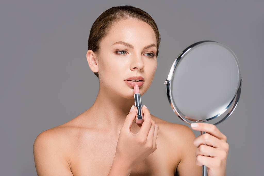 Porträt einer jungen Frau, die in den Spiegel schaut, während sie nackten Lippenstift isoliert auf Grau aufträgt - Foto, Bild