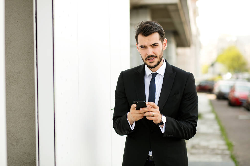 krásný elegantní muž v obleku pomocí jeho mobil a usmívá se, reciving dobré zprávy - Fotografie, Obrázek