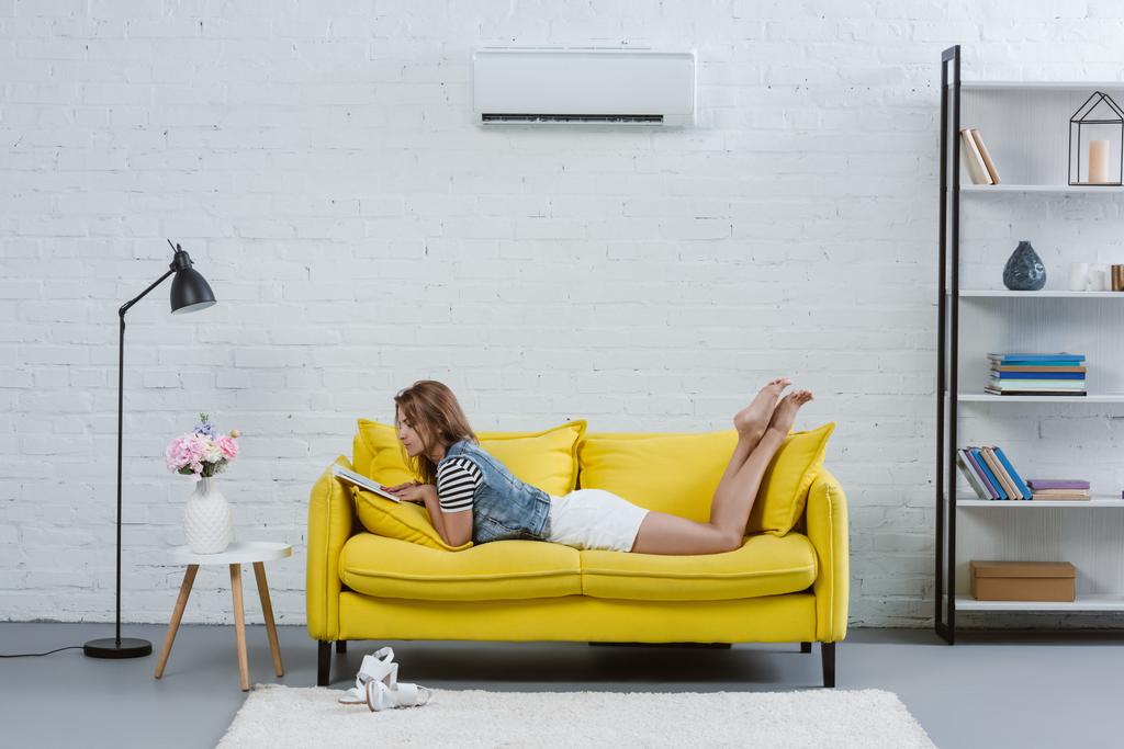 όμορφη νεαρή γυναίκα διαβάζοντας το βιβλίο στον καναπέ κάτω από το κλιματιστικό που κρέμονται στον τοίχο - Φωτογραφία, εικόνα