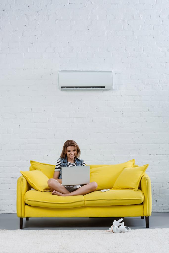 attraktive junge Frau arbeitet mit Laptop, während sie auf der Couch unter Klimaanlage an der Wand hängt - Foto, Bild