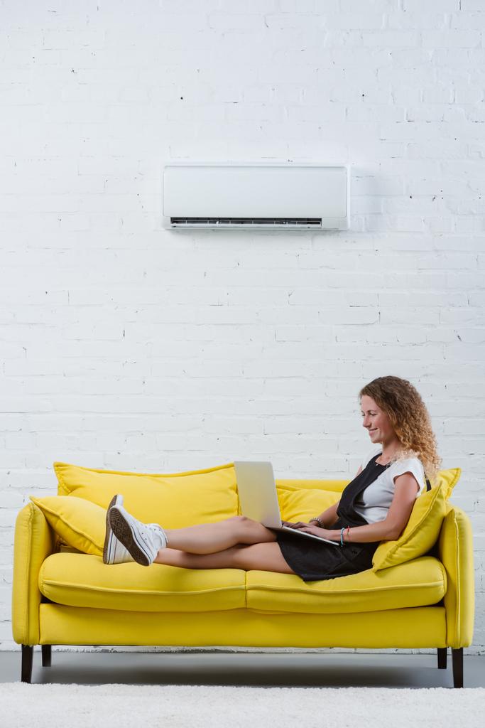 šťastná mladá žena pracující s přenosným počítačem zatímco sedí na gauči pod klimatizaci visí na zdi - Fotografie, Obrázek