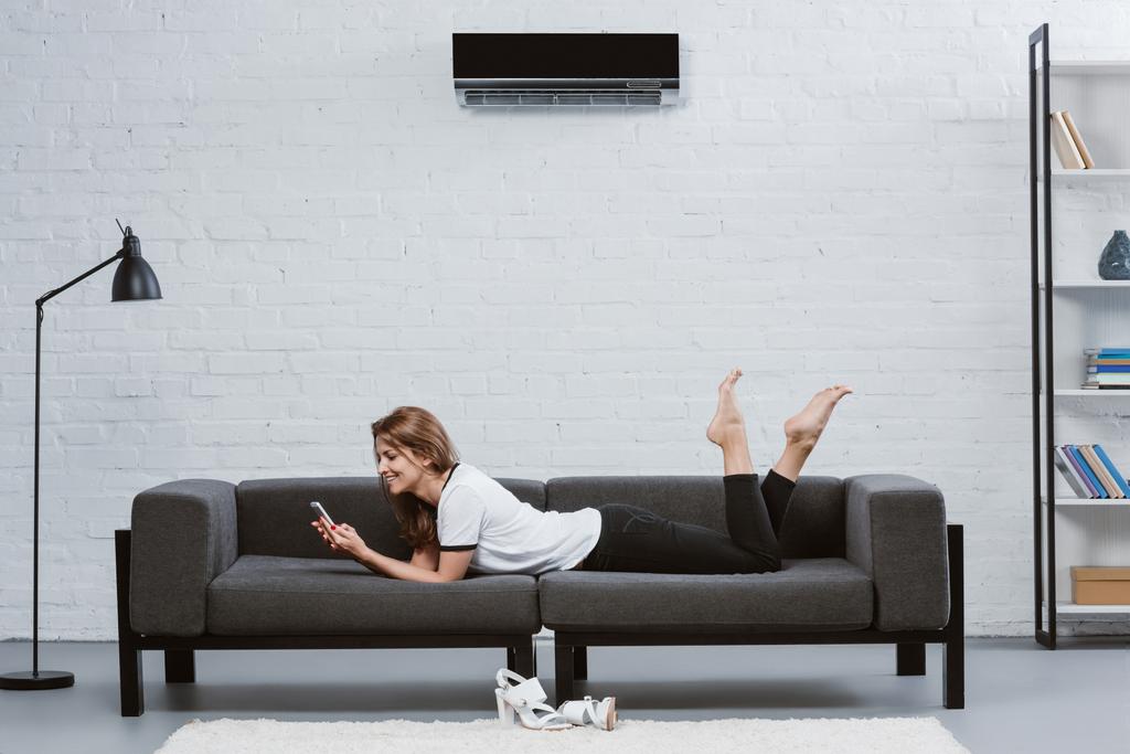 ευτυχισμένη γυναίκα χρησιμοποιώντας smartphone ξαπλωμένοι στον καναπέ κάτω από το κλιματιστικό που κρέμονται στον τοίχο - Φωτογραφία, εικόνα