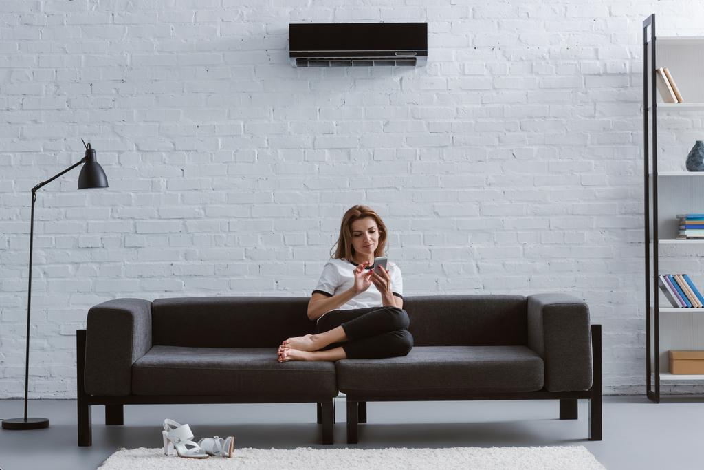 relaxed young woman using smartphone on sofa under air conditioner hanging on wall - Photo, Image