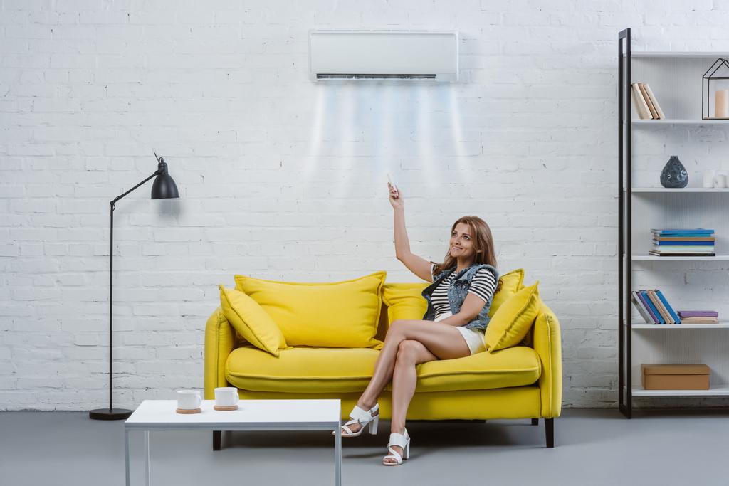 attraktive junge Frau zeigt mit Fernbedienung auf Klimaanlage, die an der Wand hängt - Foto, Bild