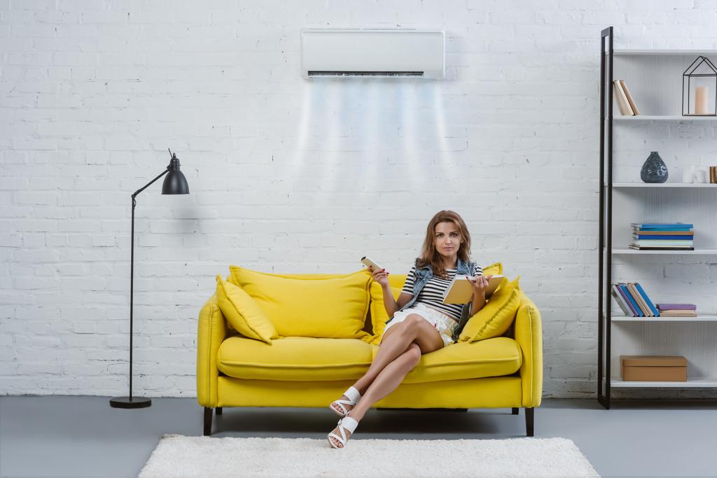 hermosa mujer joven con libro sentado en el sofá y apuntando al aire acondicionado colgando en la pared con control remoto
 - Foto, imagen