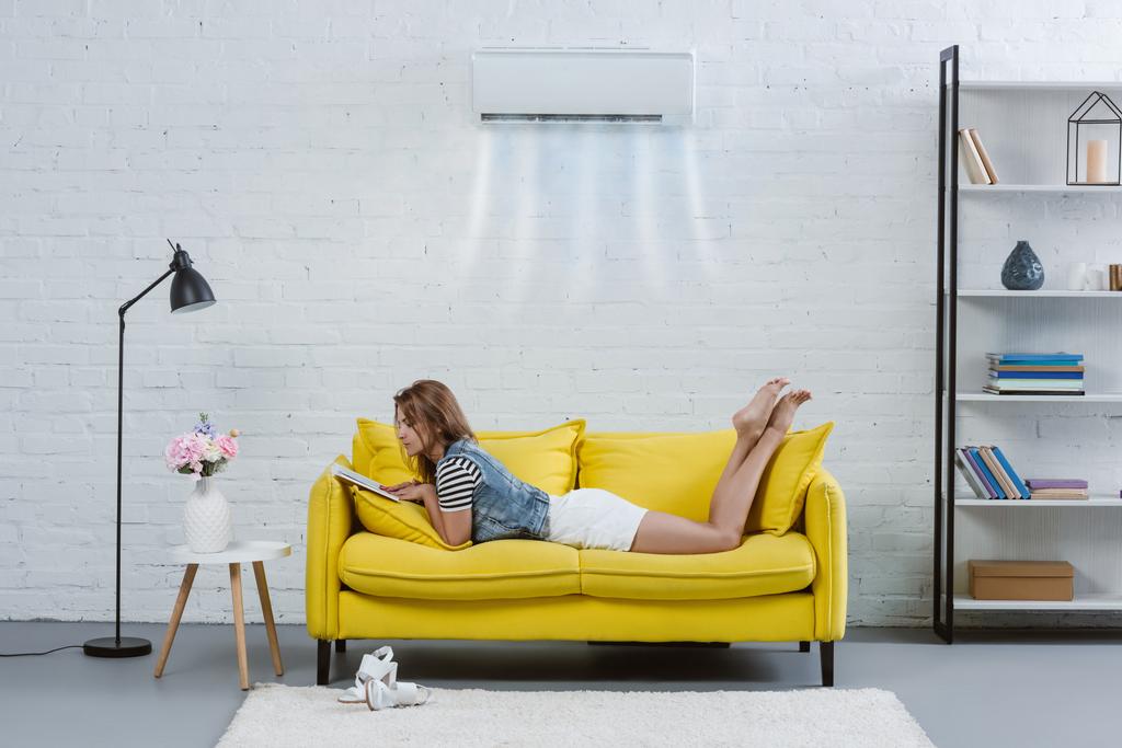 привлекательная молодая женщина читает книгу на диване под кондиционером висит на стене
 - Фото, изображение