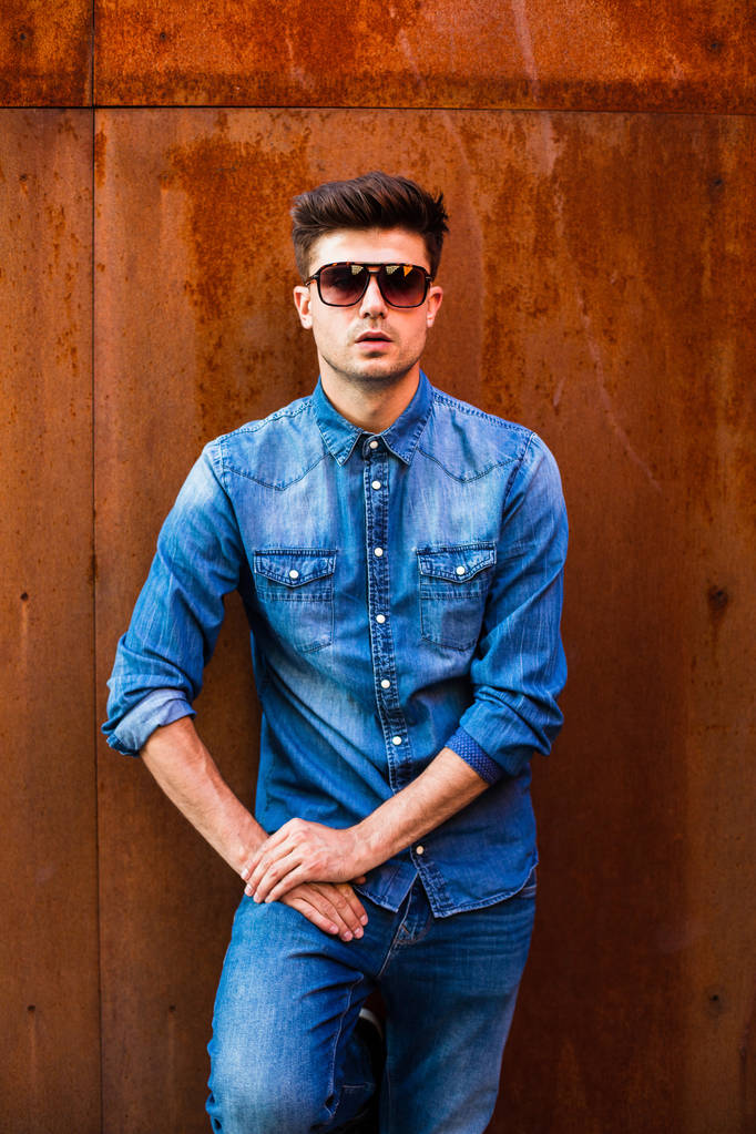 tipo seguro, hombre fresco en traje de jeans con gafas de sol de pie contra una pared oxidada
 - Foto, imagen
