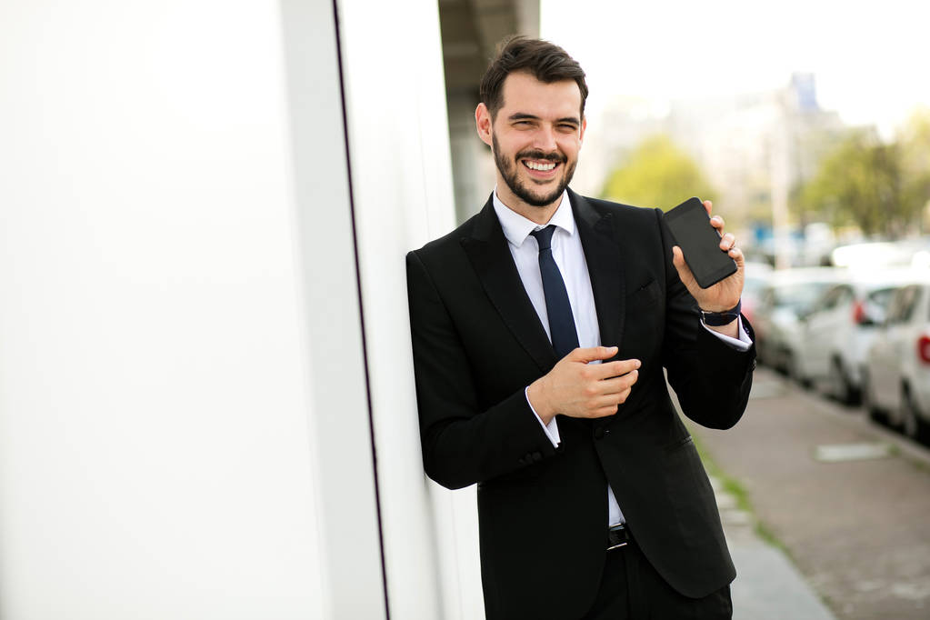 hombre elegante guapo en traje sonriendo a la cámara y mostrando su teléfono celular, afuera
 - Foto, Imagen