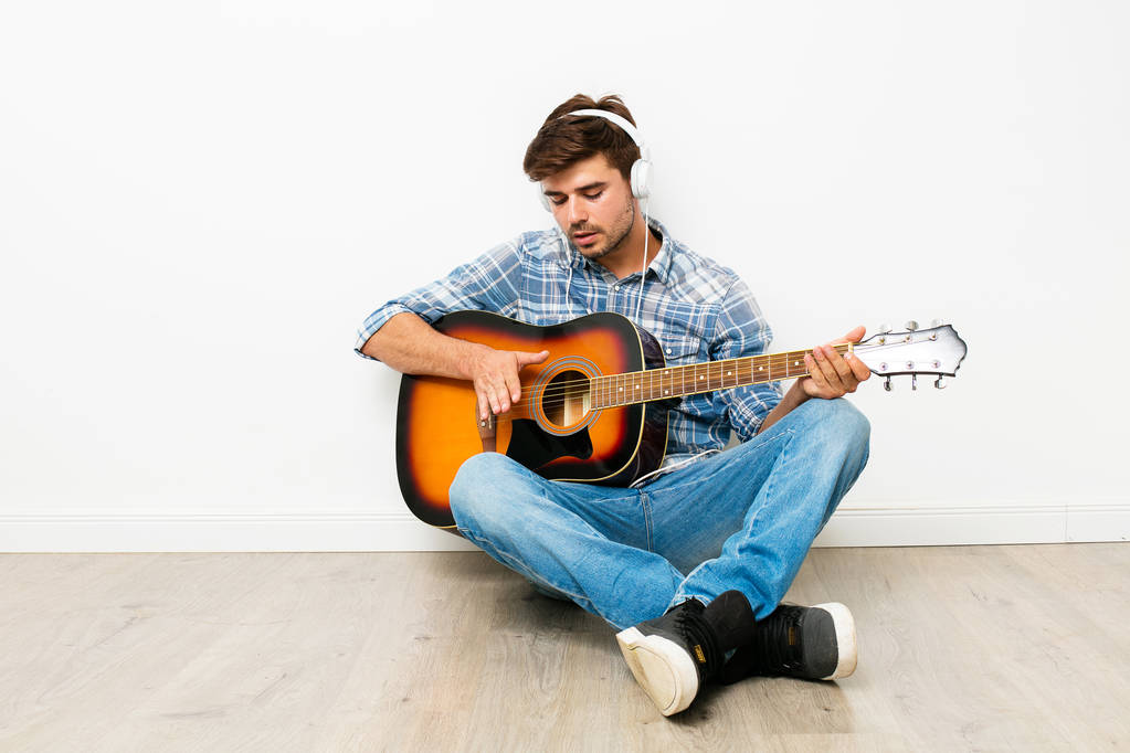 χόμπι για τα αγόρια. ο άνθρωπος παίζει κιθάρα με ακουστικά στο κεφάλι του, κάθεται στο πάτωμα, απολαμβάνοντας τον ελεύθερο χρόνο του στο δικό του σπίτι, να κάνει ό, τι θέλει, σε λευκό φόντο - Φωτογραφία, εικόνα