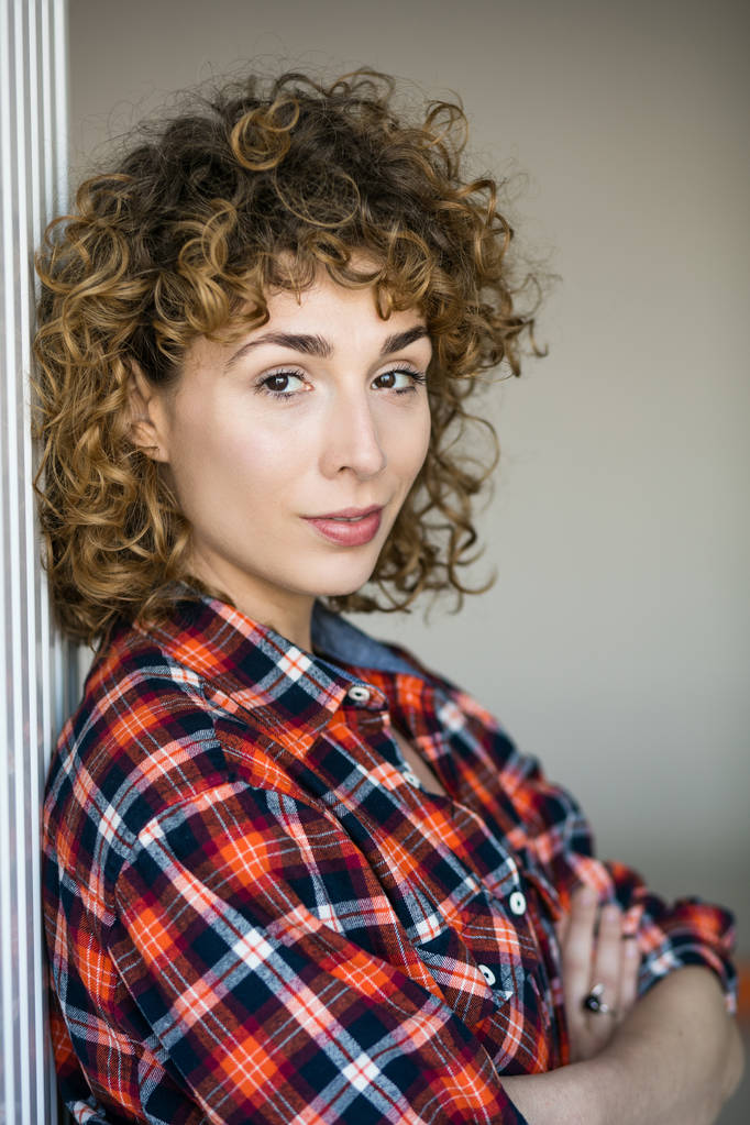 young natural curly woman smiling to camera casual dressed in a checkered shirt inside a house - Photo, Image