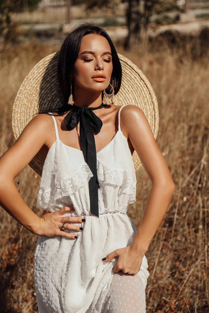 Fashion zomer buiten foto van mooie sexy vrouw met donker haar in elegante jurk met luxe strooien hoed en tas poseren in veld met droog gras - Foto, afbeelding