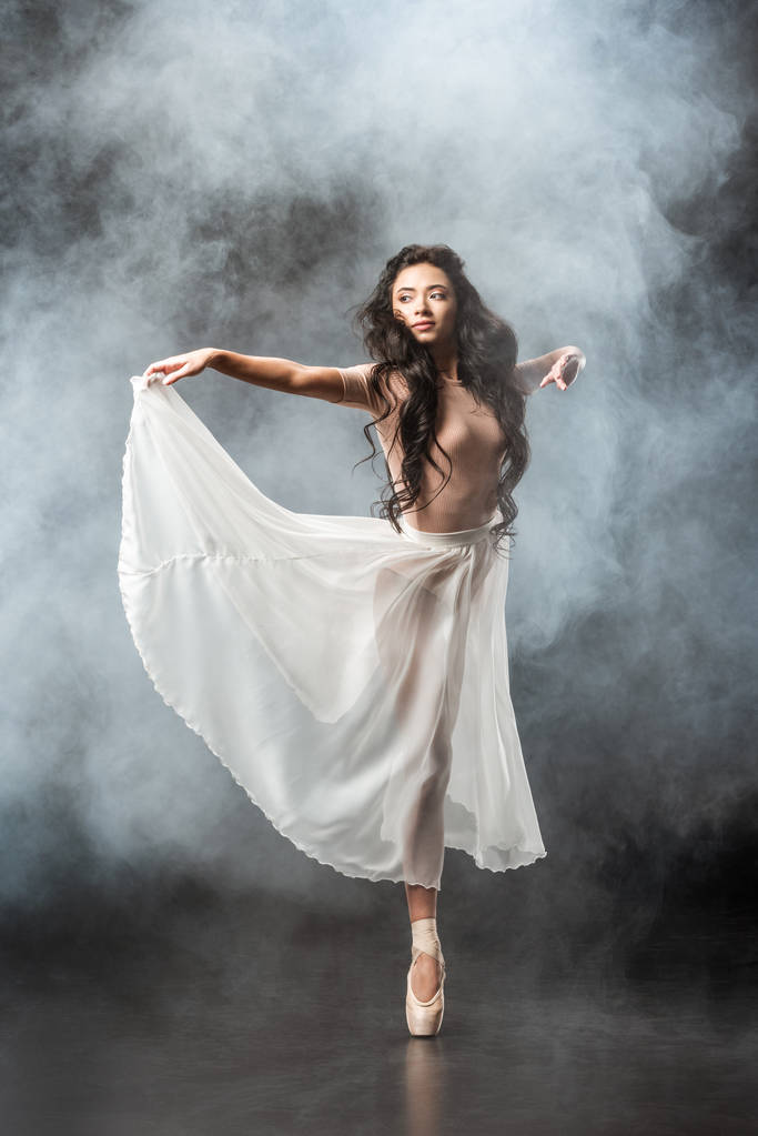 красива молода балерина в білій спідниці танцює на темному фоні з димом навколо
 - Фото, зображення