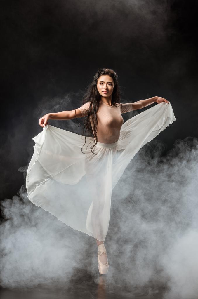 красива молода балерина в білій спідниці танцює на темному фоні з димом навколо
 - Фото, зображення