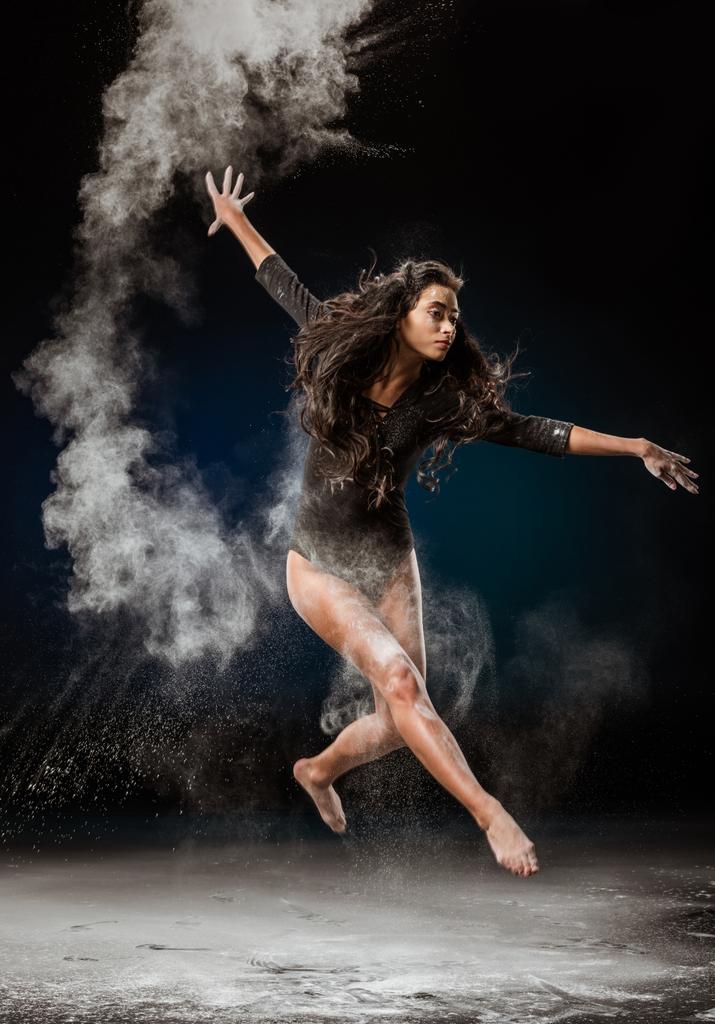 красивая балерина в черном купальнике прыгает на темном фоне с тальковым порошком вокруг
 - Фото, изображение