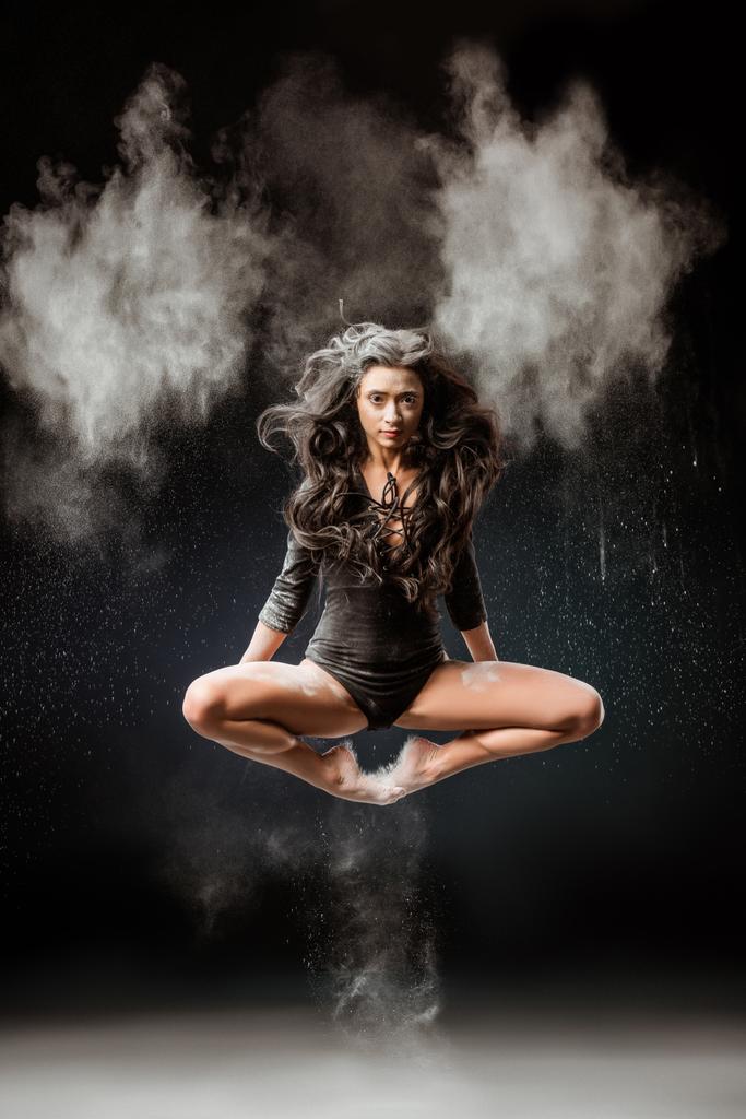 красивая балерина в черном купальнике прыгает на темном фоне с тальковым порошком вокруг
 - Фото, изображение