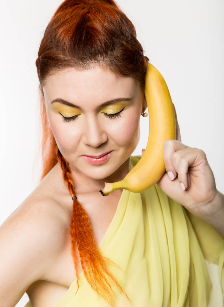 Jeune rousse femme tenant banane près de son visage comme un smartphone sur un fond blanc
. - Photo, image