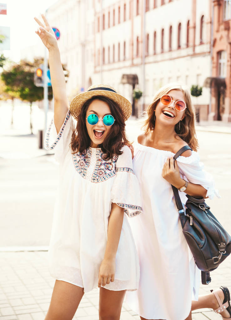 Fashion portrait of two young stylish hippie brunette and blond women models in summer sunny day in white hipster clothes posing on the street background. Going crazy - Photo, Image