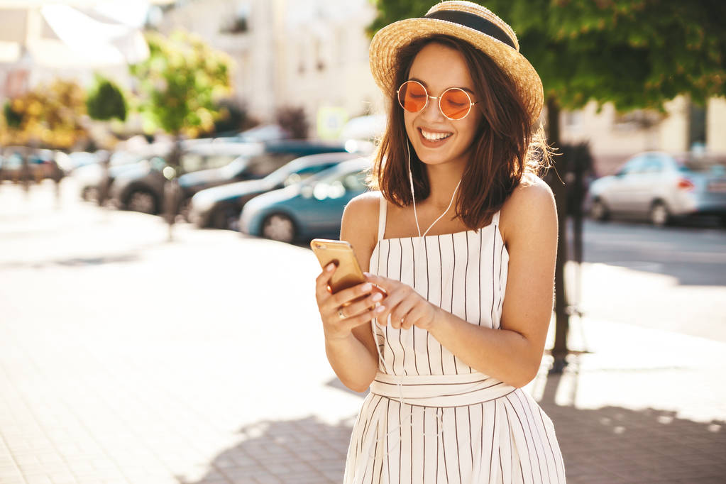 Portret van gelukkig lachend tiener meisje in zomer hipster kleding luisteren muziek op haar koptelefoon van smartphone in de straat op een zonnige dag - Foto, afbeelding