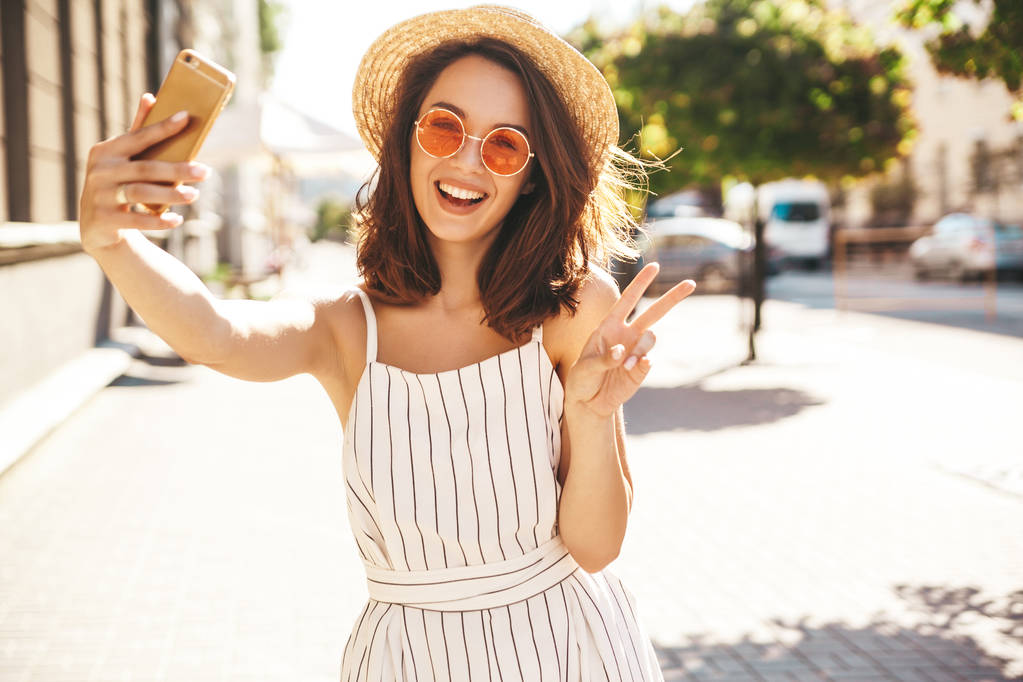 Adolescente souriante en été vêtements hipster blanc en prenant selfie sur smartphone sur fond chaud coucher de soleil. Montrer le signe de paix
 - Photo, image