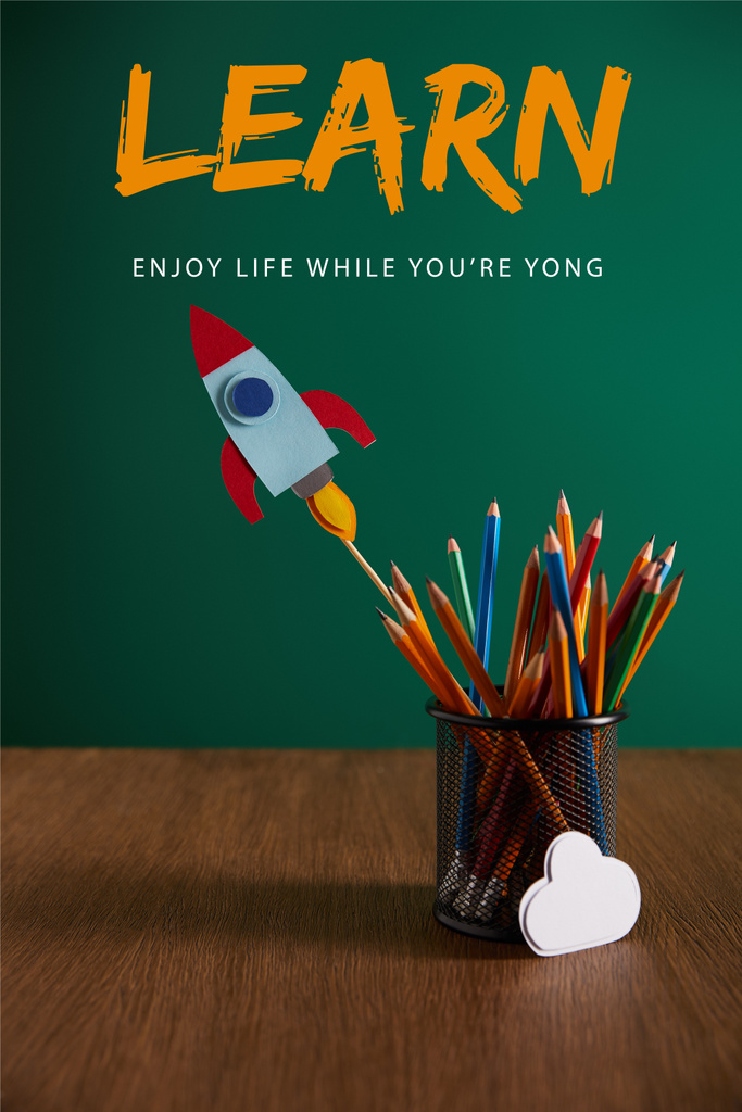 colorful pencils, rocket, cloud sign on wooden table with chalkboard on background with "learn - enjoy life while you are young" lettering - Photo, Image