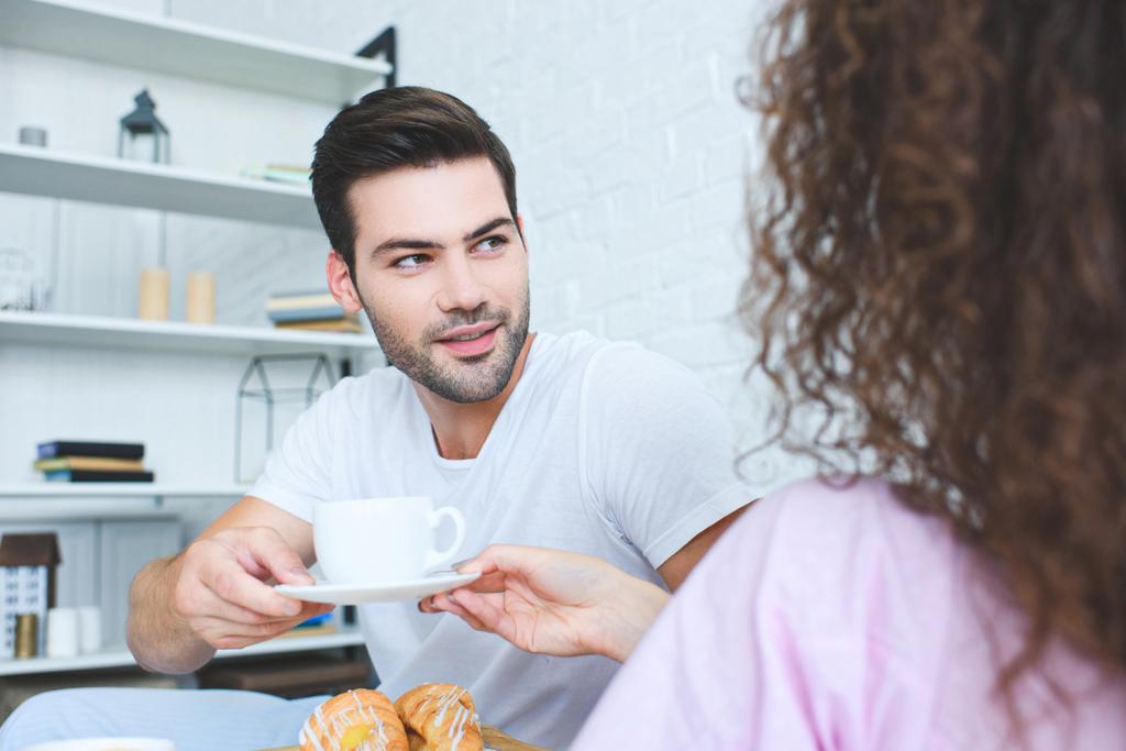 cropped shot of handsome young man holding cup of coffee and looking at girlfriend in  bedroom - Photo, Image