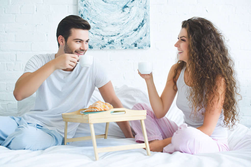 boldog fiatal pár pizsamában kávét iszik, és mosolyogva egymást, miközben ül ágyas - Fotó, kép