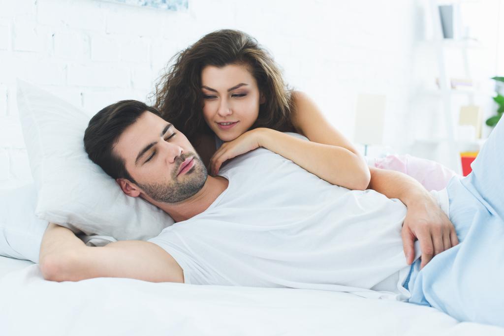 hermosa sonriente joven mujer mirando durmiendo novio en dormitorio
 - Foto, imagen