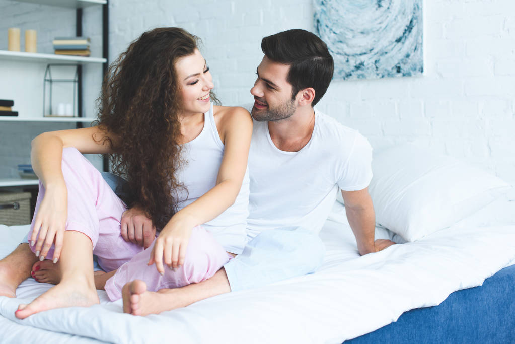 красива щаслива молода пара в піжамі сидить на ліжку і посміхається один одному
 - Фото, зображення