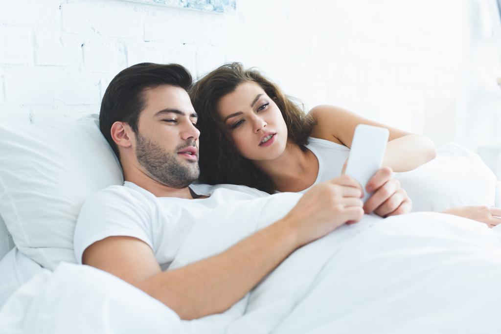 νεαρό ζευγάρι ξαπλωμένη στο κρεβάτι και χρησιμοποιώντας smartphone μαζί - Φωτογραφία, εικόνα