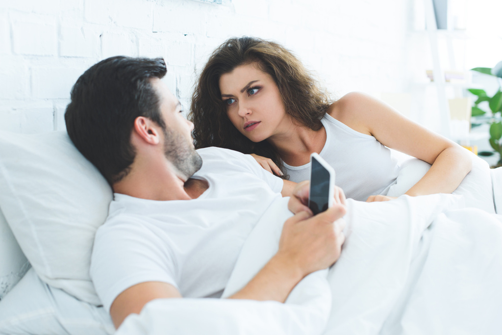 Junges Paar schaut einander an und streitet sich im Bett über Smartphone, Beziehungsschwierigkeiten - Foto, Bild