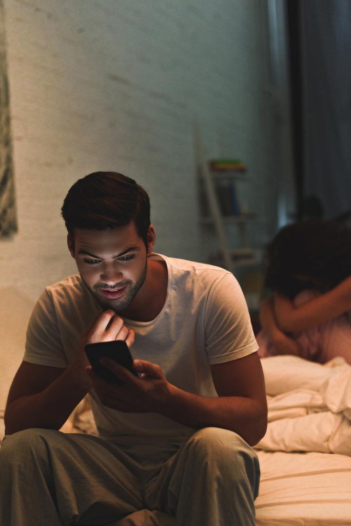 Junger Mann nutzt Smartphone, während aufgebrachte Freundin im Bett sitzt, Beziehungsschwierigkeiten  - Foto, Bild
