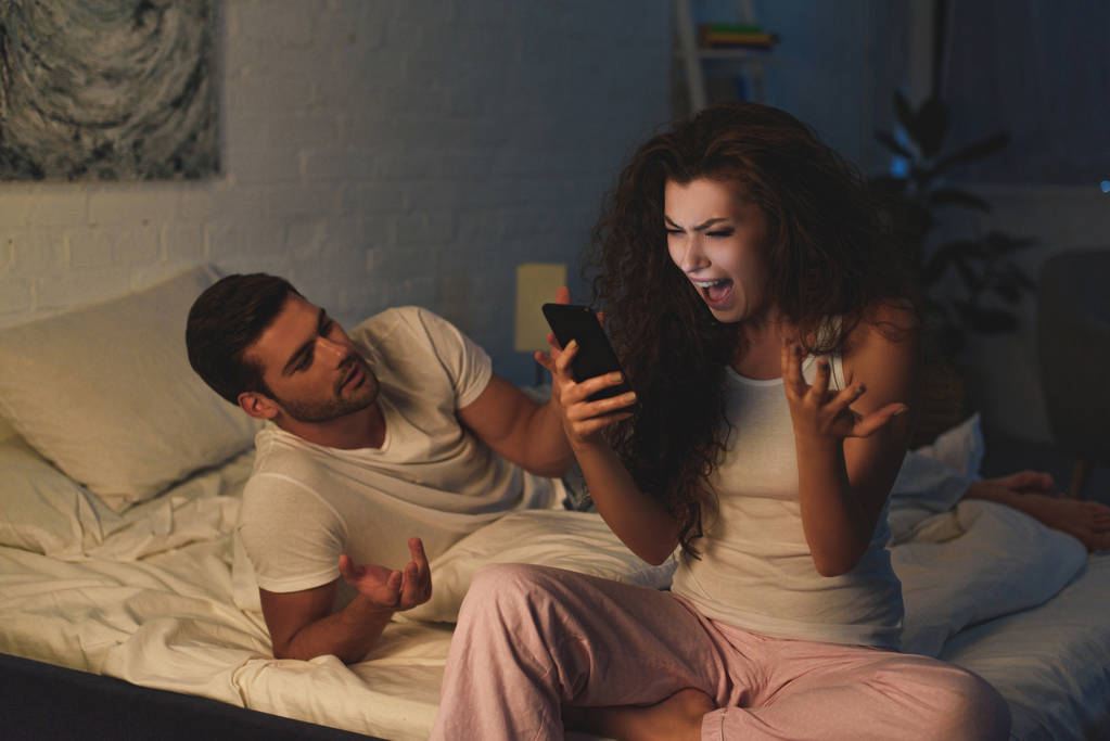 θυμωμένος νεαρή γυναίκα χρησιμοποιώντας smartphone και ουρλιάζοντας ενώ ο φίλος ξαπλωμένος στο κρεβάτι τη νύχτα - Φωτογραφία, εικόνα