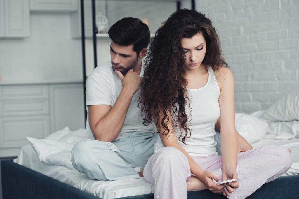 jonge vrouw met behulp van smartphone terwijl boos man zit achter op bed, relatie problemen concept - Foto, afbeelding