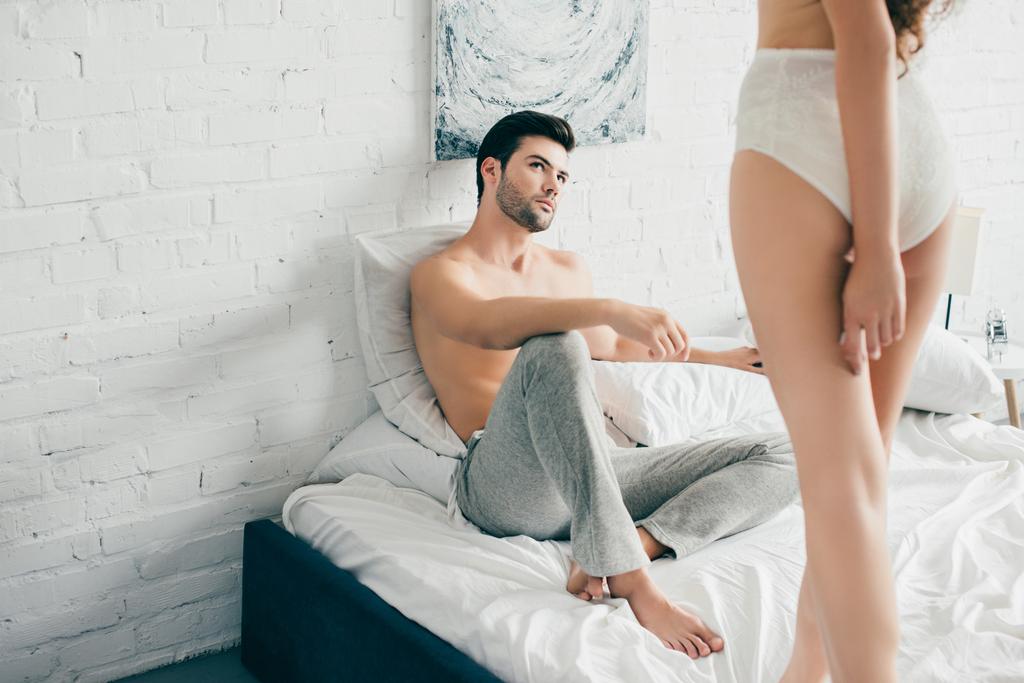 обрезанный снимок красивого мужчины, смотрящего на чувственную девушку в нижнем белье, стоящую на кровати
    - Фото, изображение